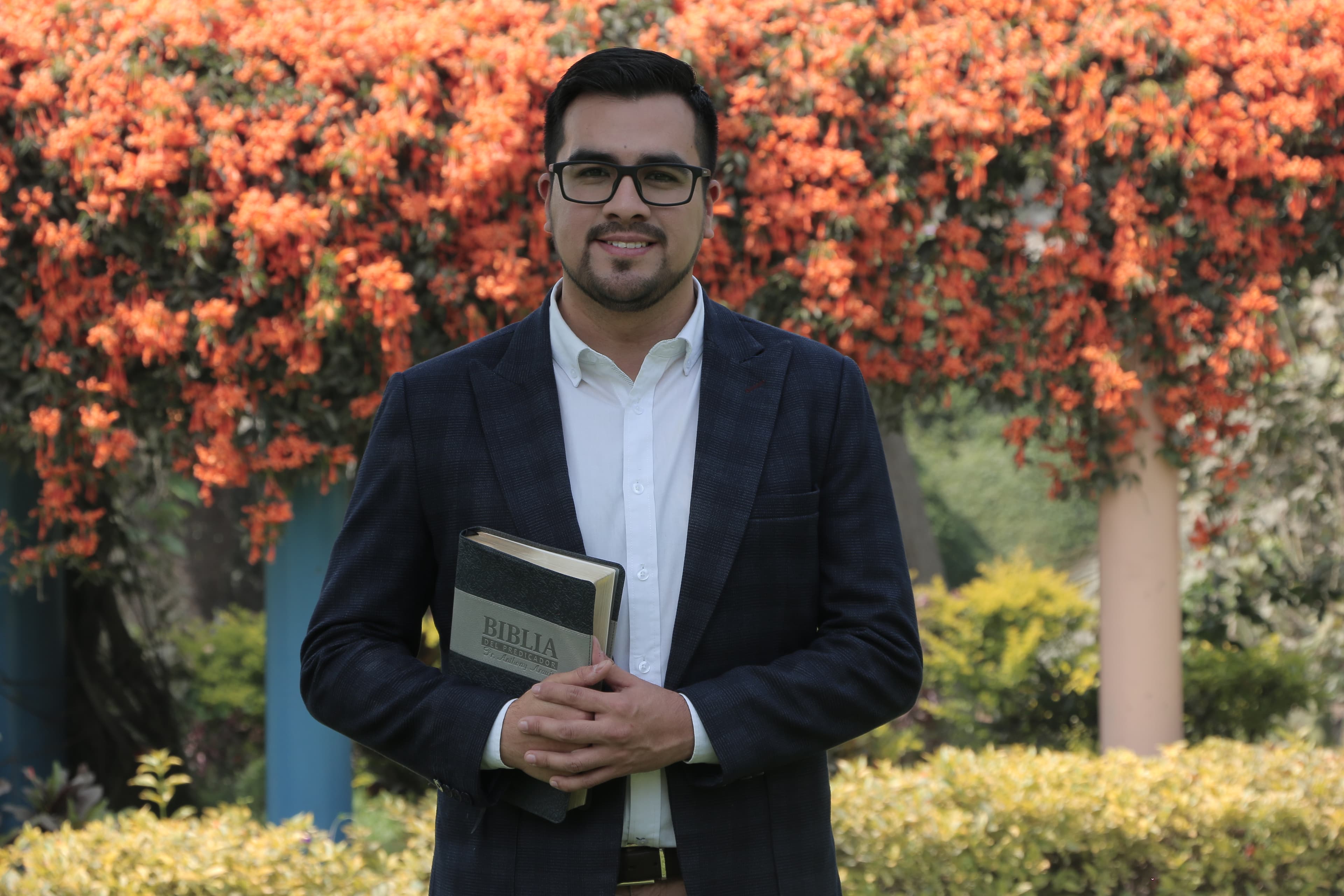 Anthony Araujo al aire libre, sosteniendo su Biblia, frente a un árbol con flores anaranjadas.