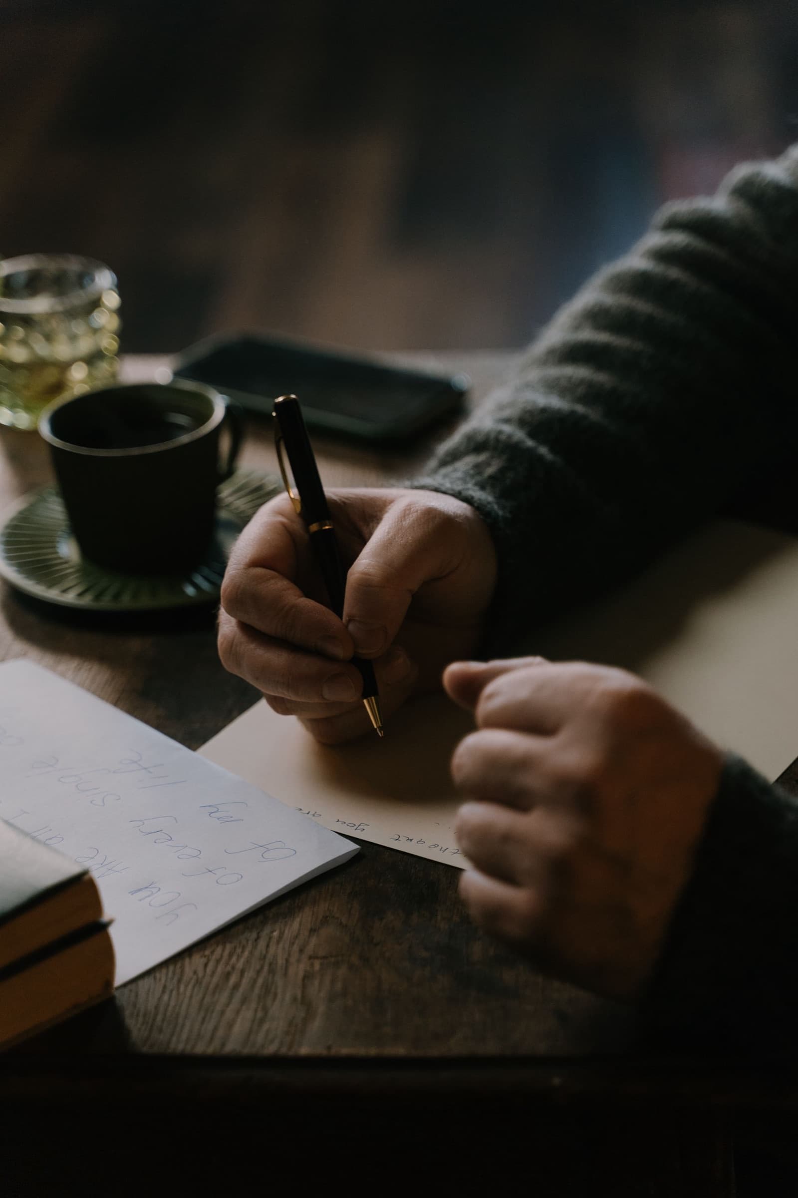 Manos escribiendo con una pluma sobre papel, junto a una taza, en un ambiente cálido de cafetería.